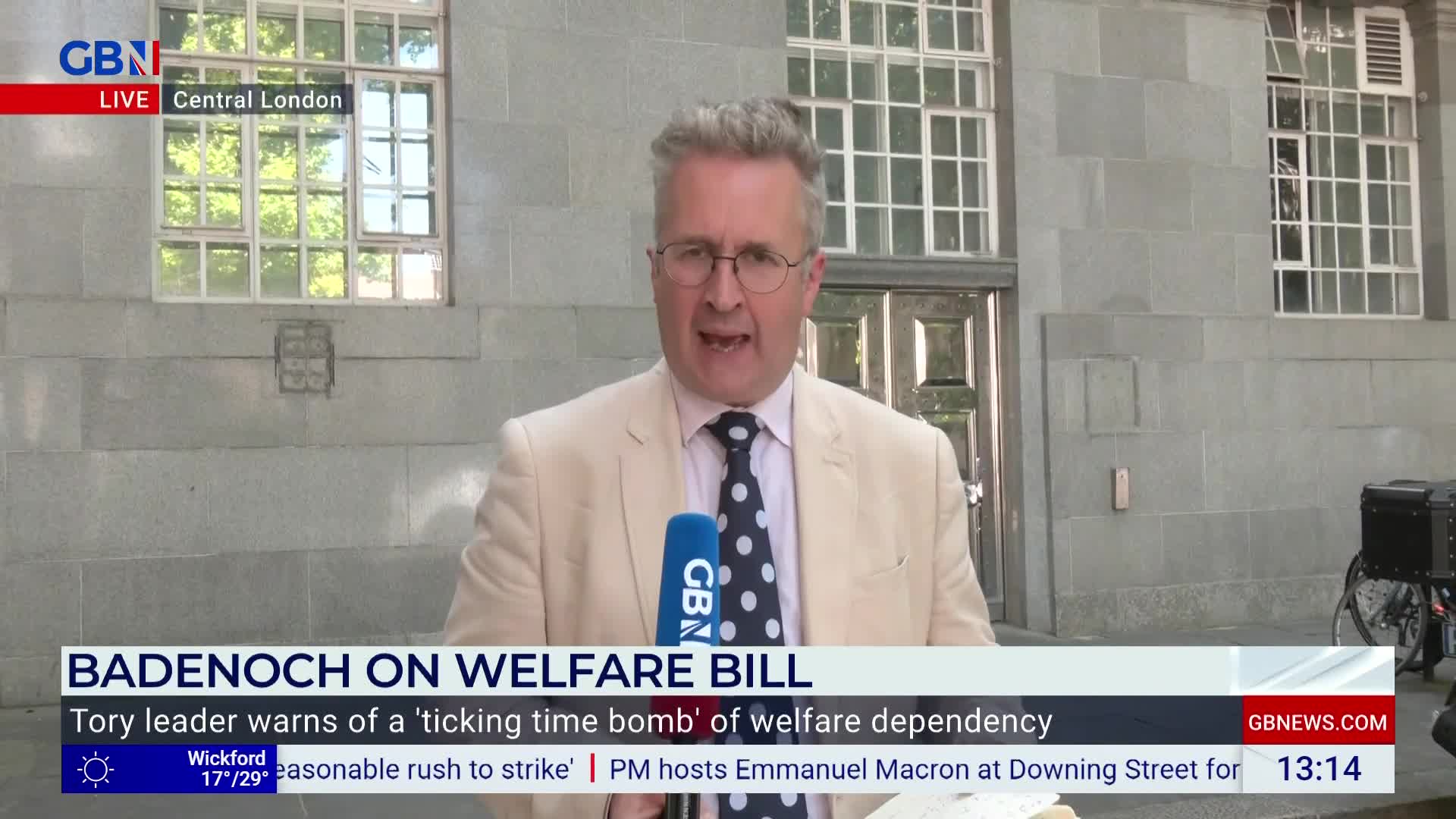 A man in a light-colored suit and polka-dot tie speaks directly into a GB News microphone in Central London. Behind him, a stone building with large windows provides a backdrop for the live broadcast.
