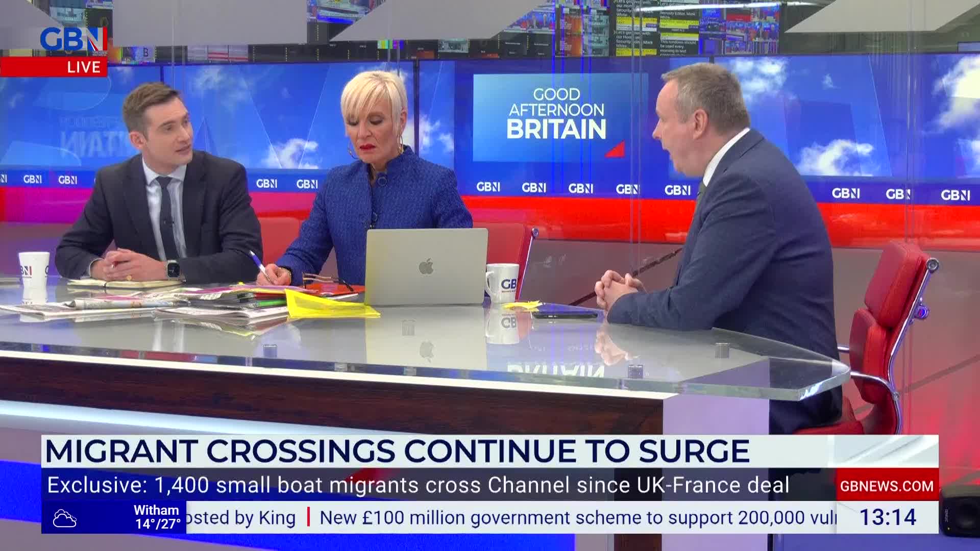 Three people sit at a desk in a GB News studio; one man on the right gestures as he speaks. The chyron at the bottom of the screen discusses migrant crossings from the UK.
