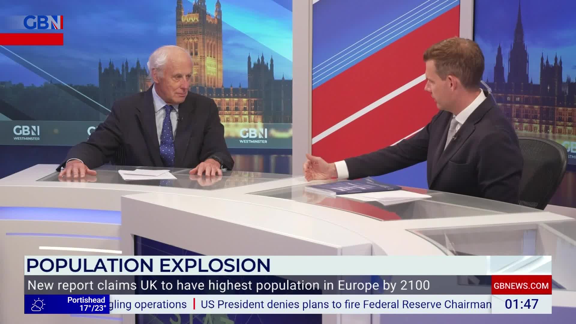 Two men in suits are seated at a desk, facing each other on a GB News set. The backdrop shows the Houses of Parliament, and the lower third of the screen displays a headline about the UK's population.
