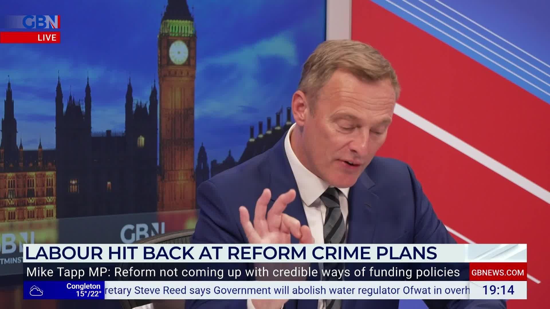 A man in a suit gestures with his hand while speaking on GB News. Behind him, a backdrop shows the Houses of Parliament in London.
