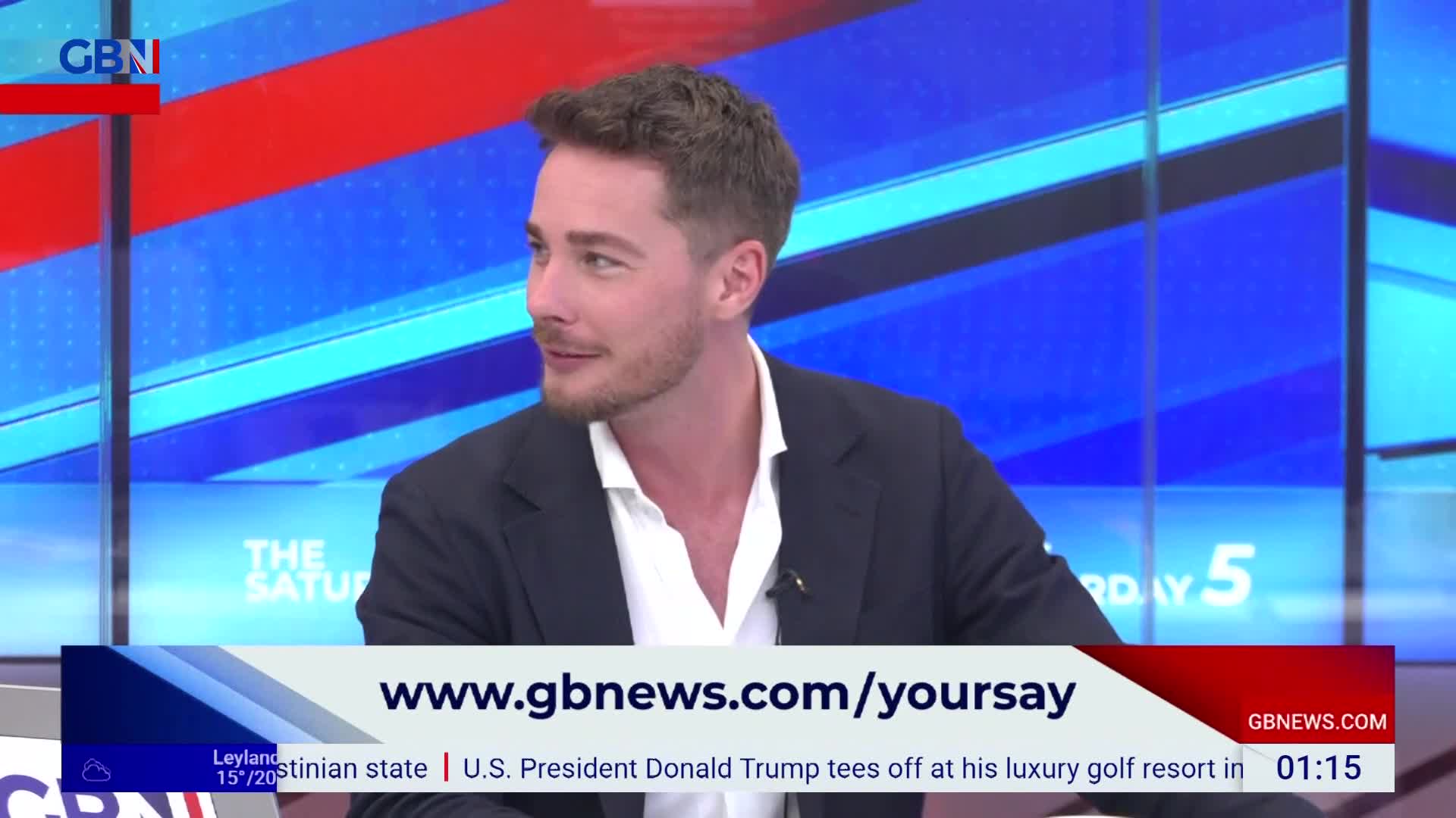 A man in a dark suit and open-collared shirt is speaking on GB News, his gaze directed off-screen.  Below him, a banner encourages viewers to visit the website, www.gbnews.com/yoursay.
