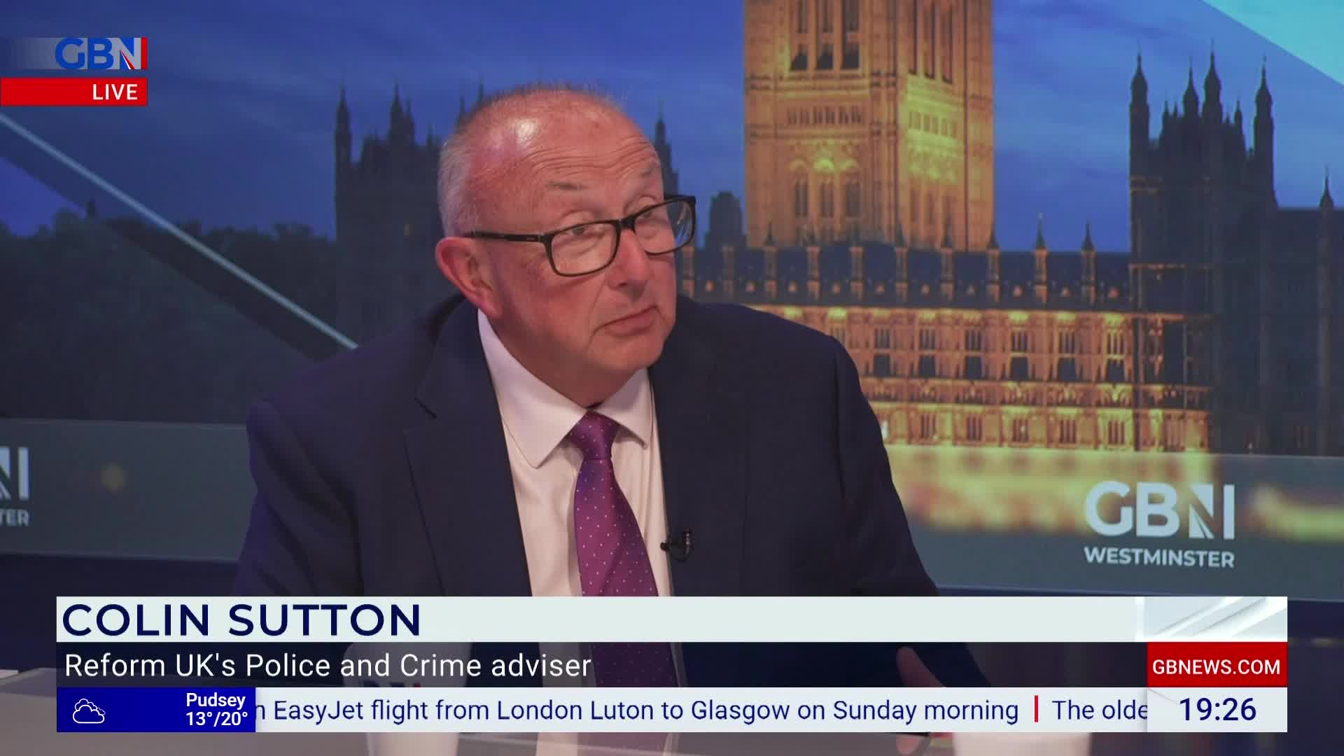 A man in a suit and tie sits at a desk, looking to his left on a GB News set. The backdrop shows the Houses of Parliament, and a lower third identifies him as Colin Sutton, a police and crime advisor.
