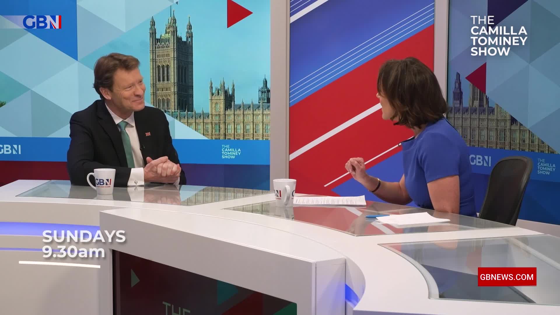 A man in a suit sits at a desk, smiling at the woman across from him on the GB News set. She gestures with a pen, likely asking a question on "The Camilla Tominey Show" broadcast from the United Kingdom.
