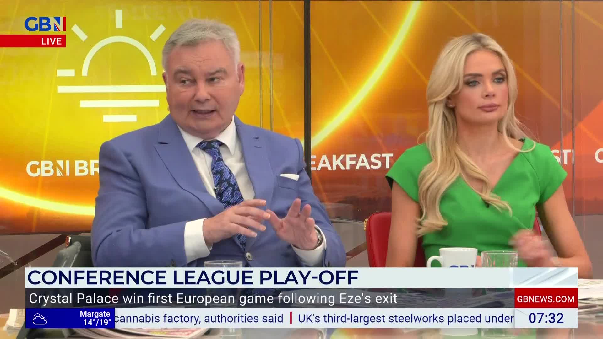The man in the blue suit gestures with his hands, speaking animatedly on GB News. Beside him, a woman in a green dress listens intently, her expression composed.
