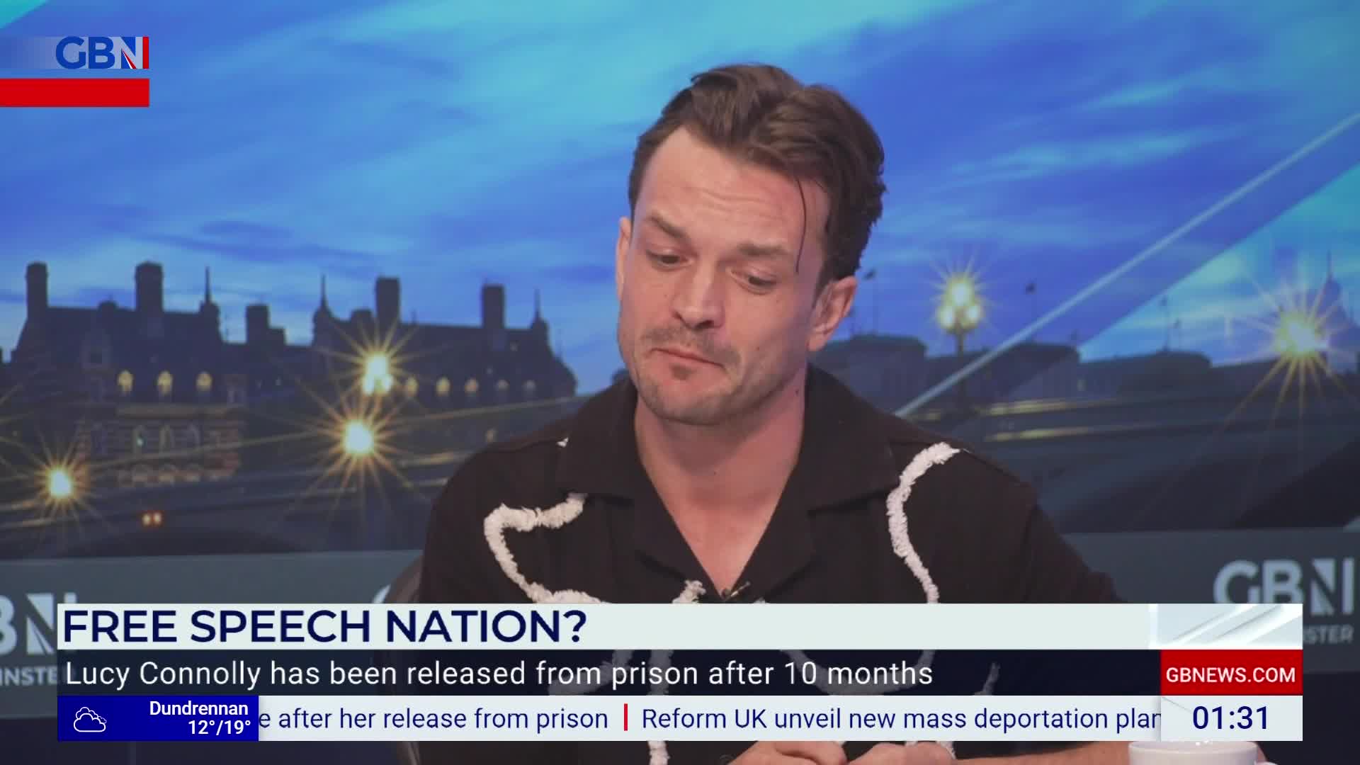 A man in a black shirt with white trim is speaking on GB News, the backdrop showing a nighttime view of a building. The lower third of the screen displays a headline about free speech and a news ticker.
