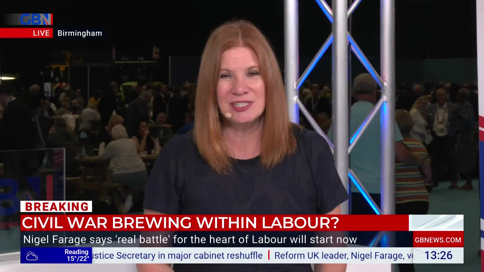 A woman with red hair is speaking on GB News, in Birmingham. Behind her, a crowd of people mill about, some seated at tables.
