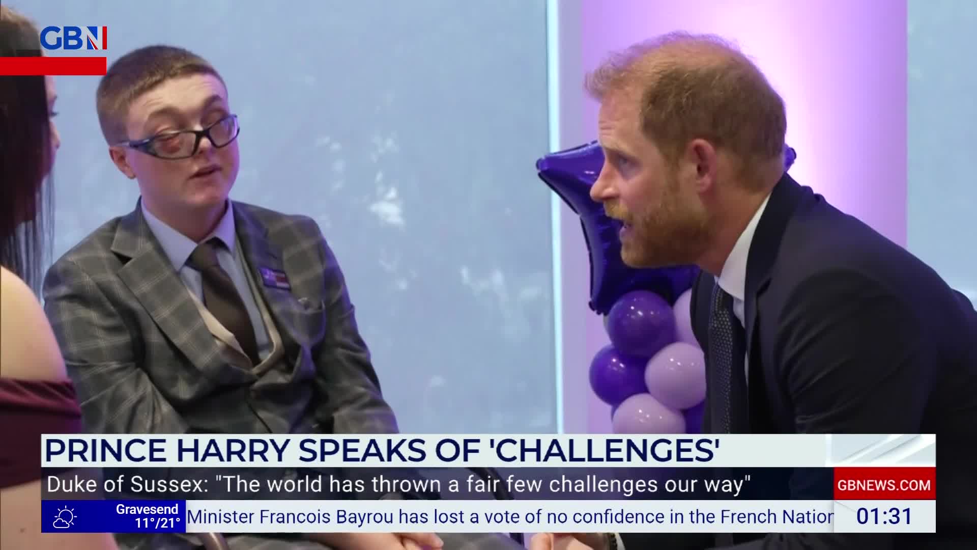 Prince Harry, on GB News, gestures animatedly, speaking to someone off-camera. The other person, wearing a suit, listens intently, framed by a backdrop of purple balloons.
