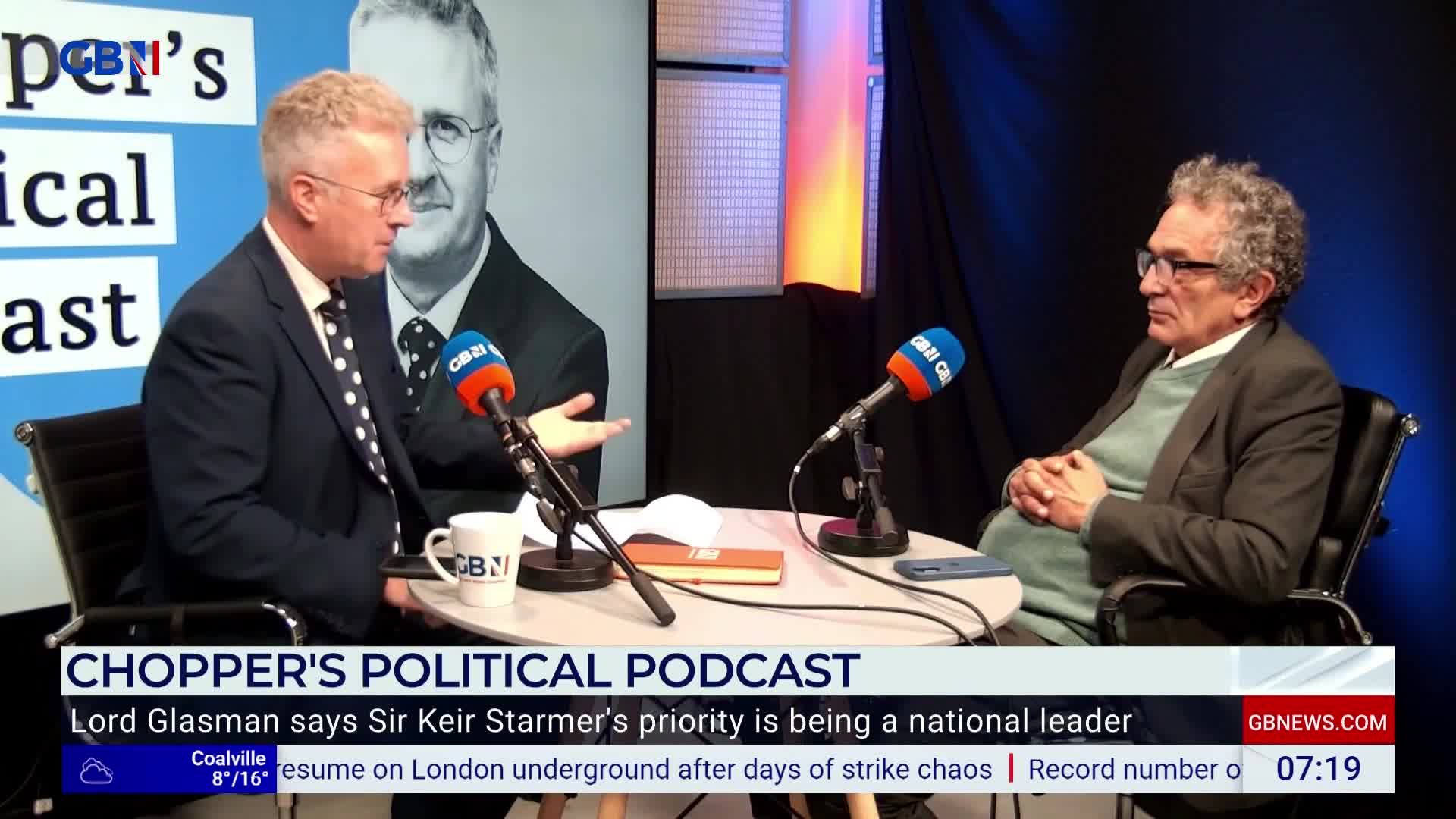 A man in a suit gestures with his right hand, speaking into a microphone on a GB News set. Across from him, another man with glasses and a sweater sits, listening intently.
