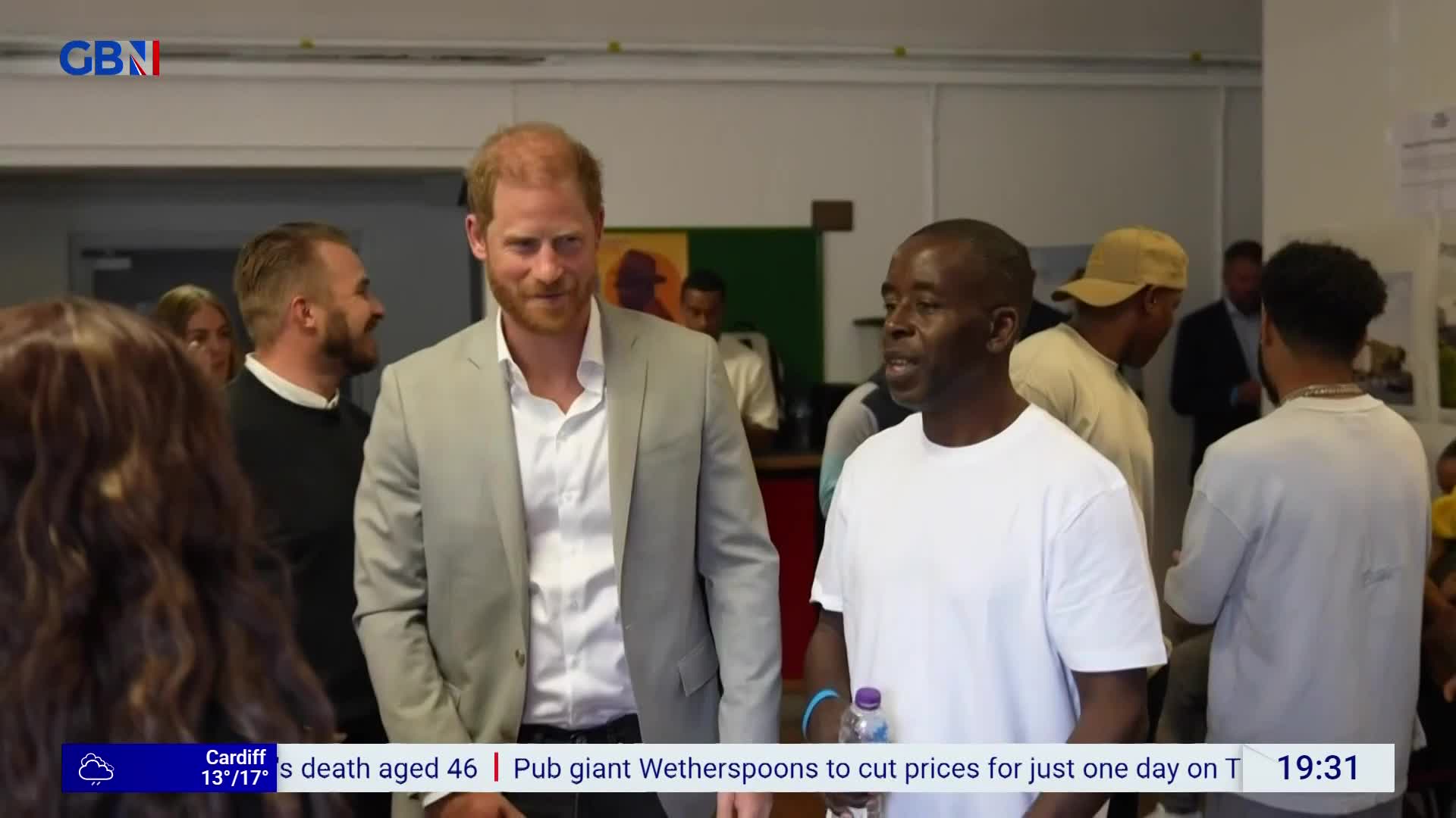 Prince Harry, in a light grey suit, is walking and looking towards the camera. A man in a white t-shirt is speaking, gesturing with his hands as he walks beside him.
