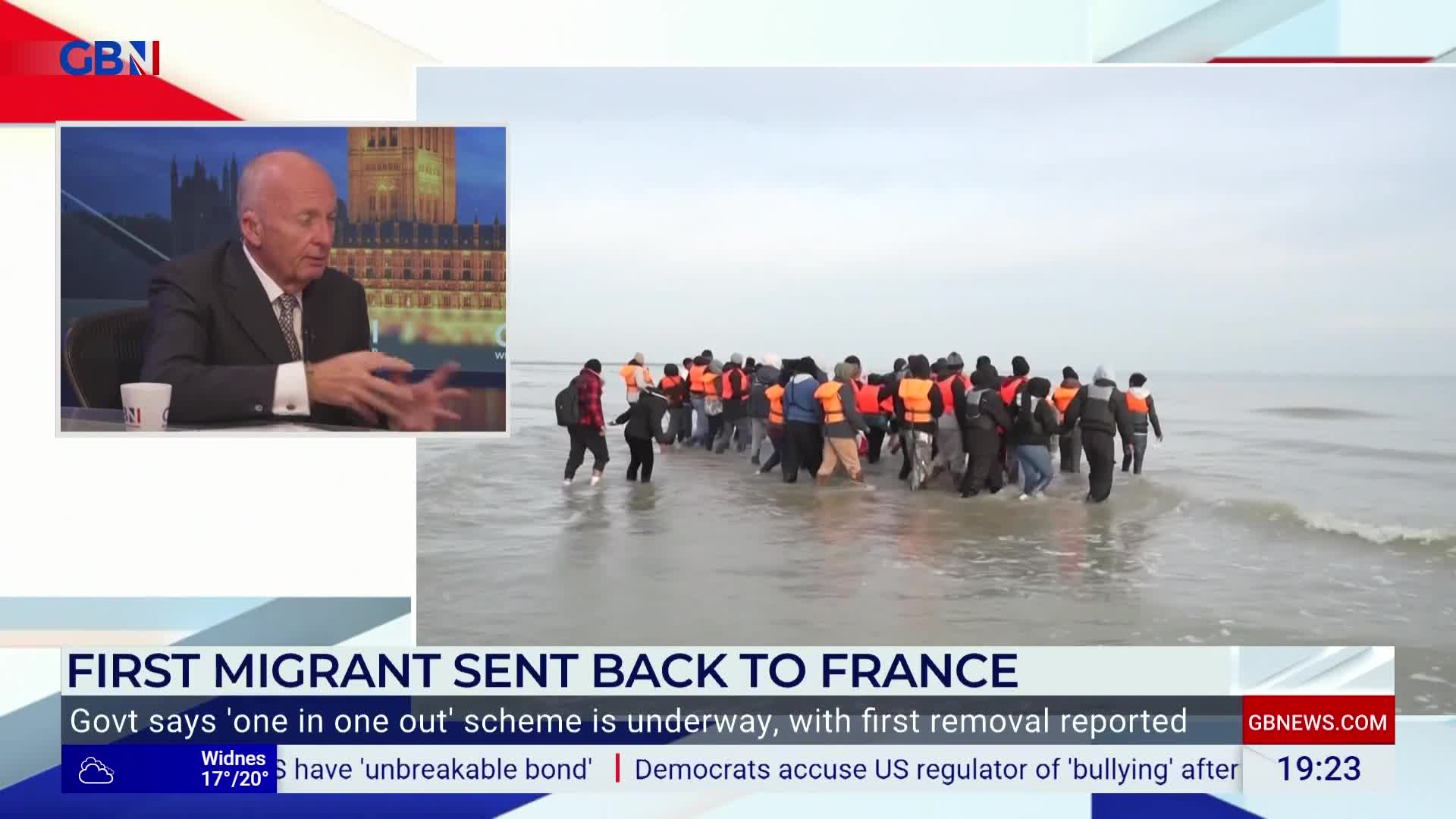 A group of people wearing orange life vests wade through shallow water. On the screen, a GB News anchor discusses the UK government's "one in one out" scheme.
