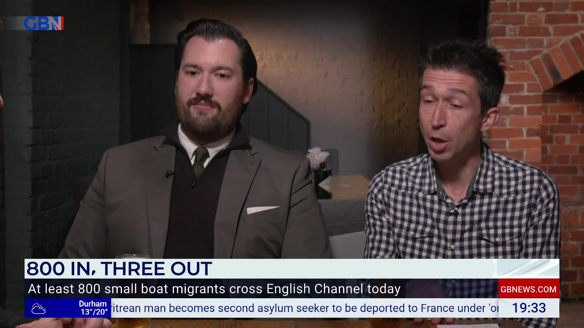 Two men are seated, presumably on a GB News set, discussing the day's news. The screen displays a headline about migrants crossing the English Channel.
