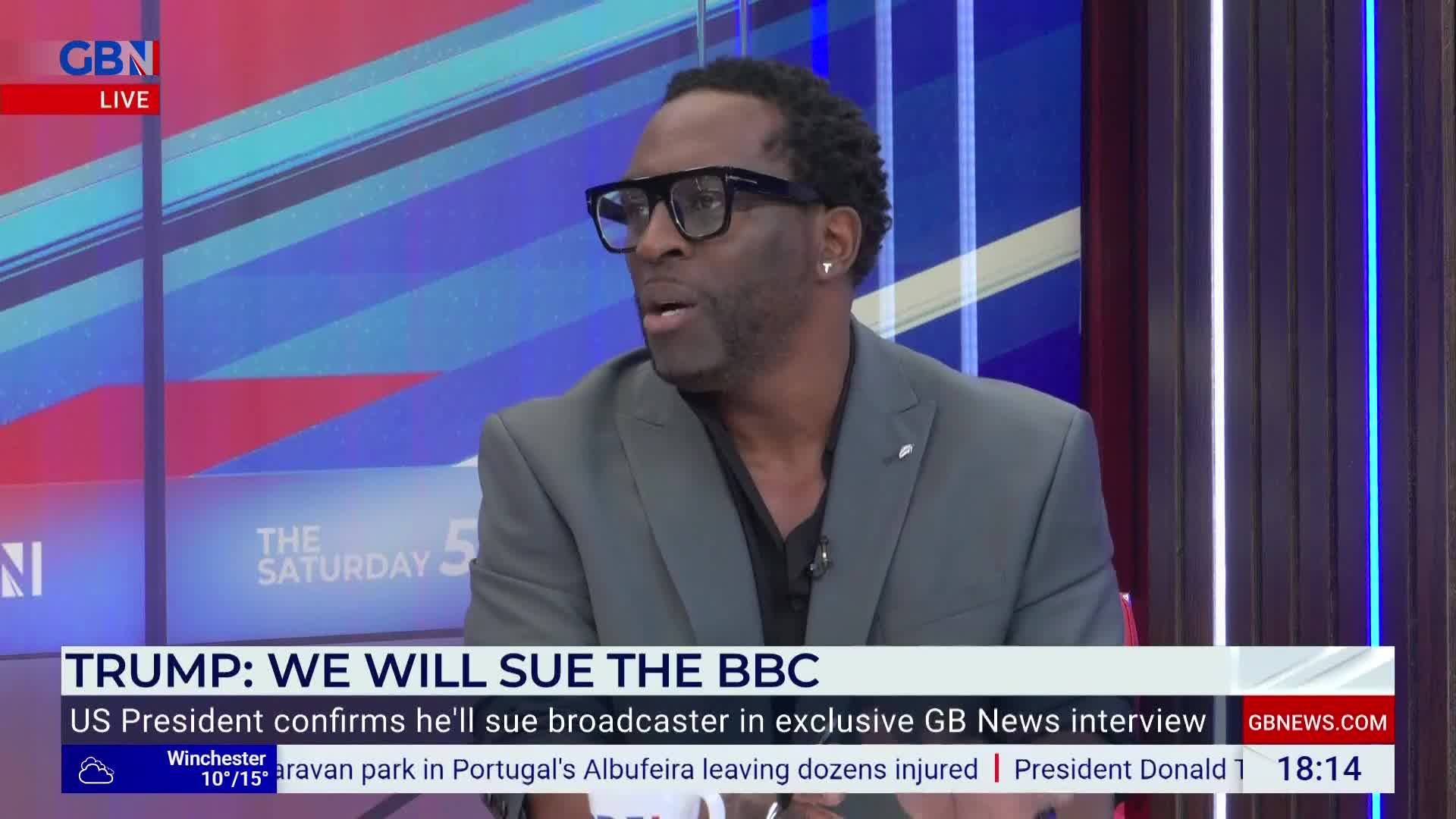 A man in a gray blazer and black glasses is speaking on the GB News set. The lower third headline announces a lawsuit against the BBC.
