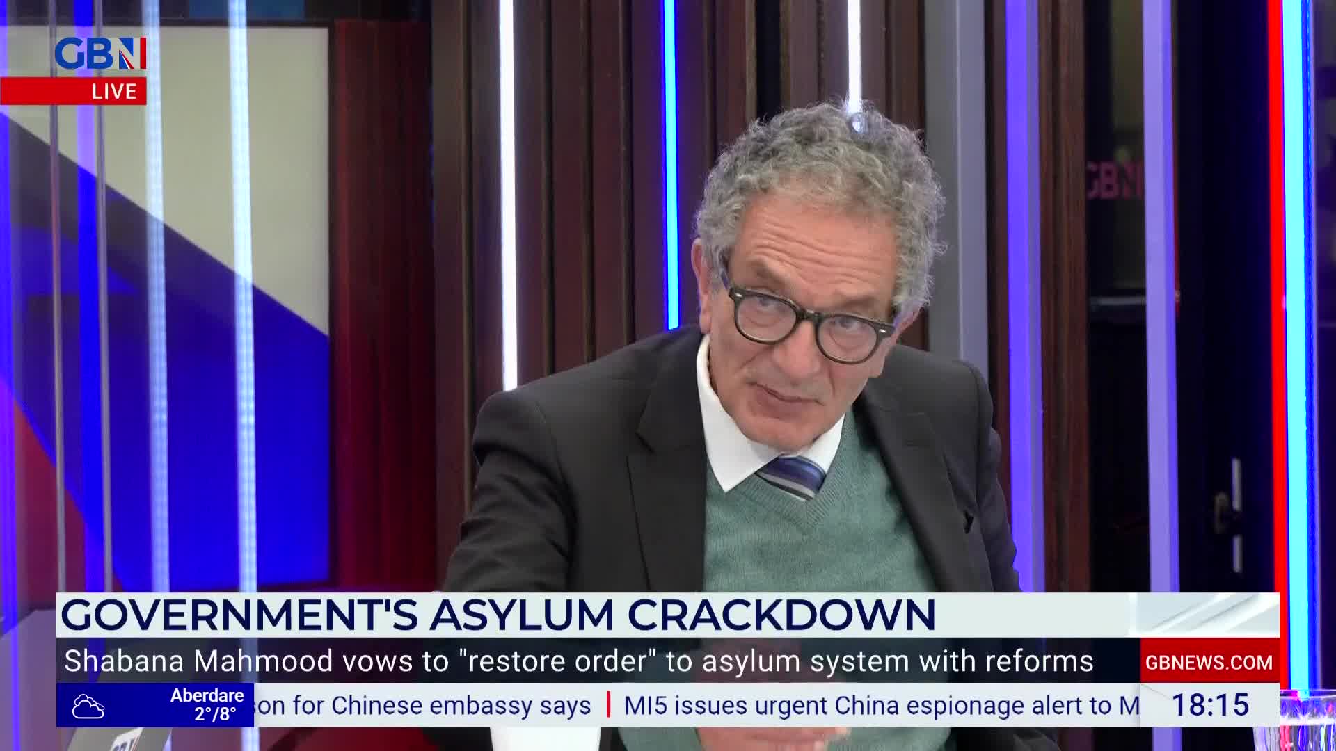 A man in glasses, wearing a suit jacket and sweater, is speaking intently on a GB News set. The headline "GOVERNMENT'S ASYLUM CRACKDOWN" appears below him.
