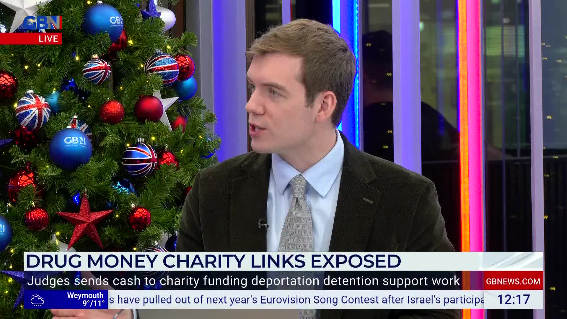 A man in a suit jacket is speaking, his gaze directed off-screen. Behind him, a Christmas tree decorated with Union Jack ornaments stands next to a screen displaying the GB News logo.
A man in a suit jacket is speaking, his gaze directed off-screen. Behind him, a Christmas tree decorated with Union Jack ornaments stands next to a screen displaying the GB News logo.