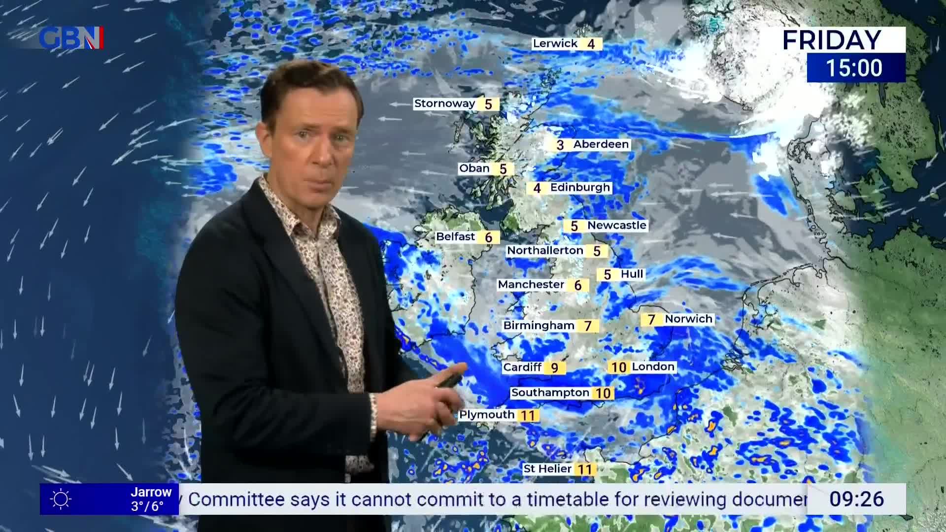 A man in a dark jacket stands before a weather map of the United Kingdom, pointing at the screen. The map shows areas of rain and cloud across the country, with temperatures displayed for various cities.