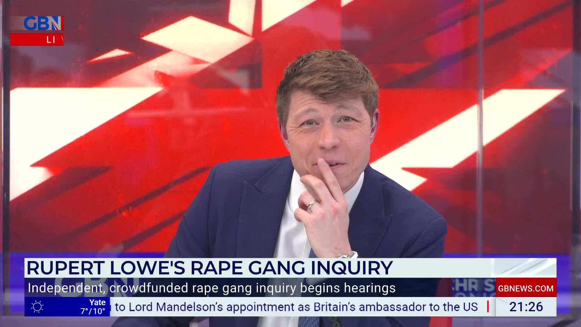 A man in a blue suit and white shirt is speaking on GB News, his fingers touching his chin. The screen behind him displays a graphic with red and white angular shapes.