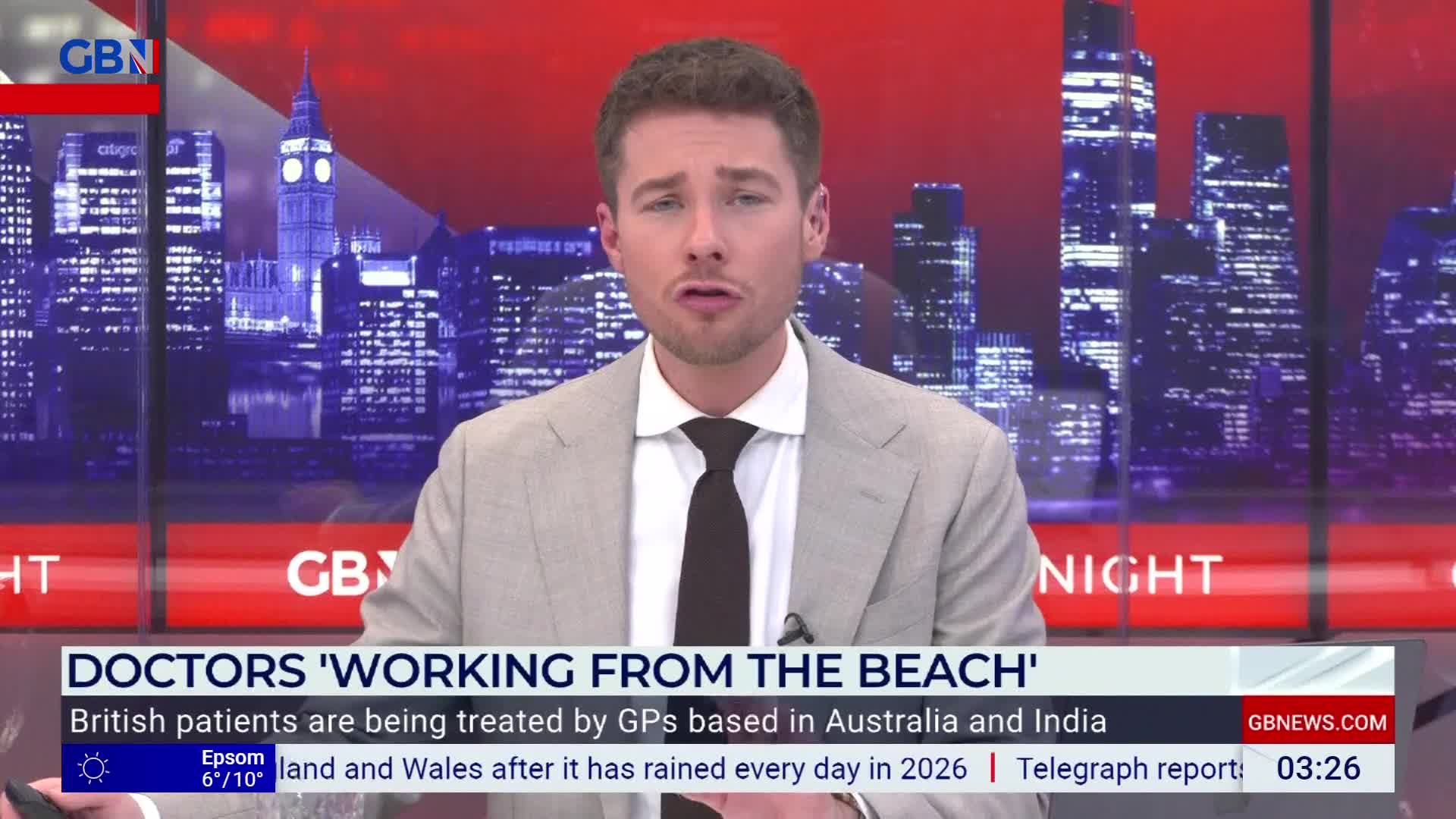A man in a suit stands before a backdrop of the London skyline. He's speaking, his expression serious, as the headline "DOCTORS 'WORKING FROM THE BEACH'" flashes on screen.