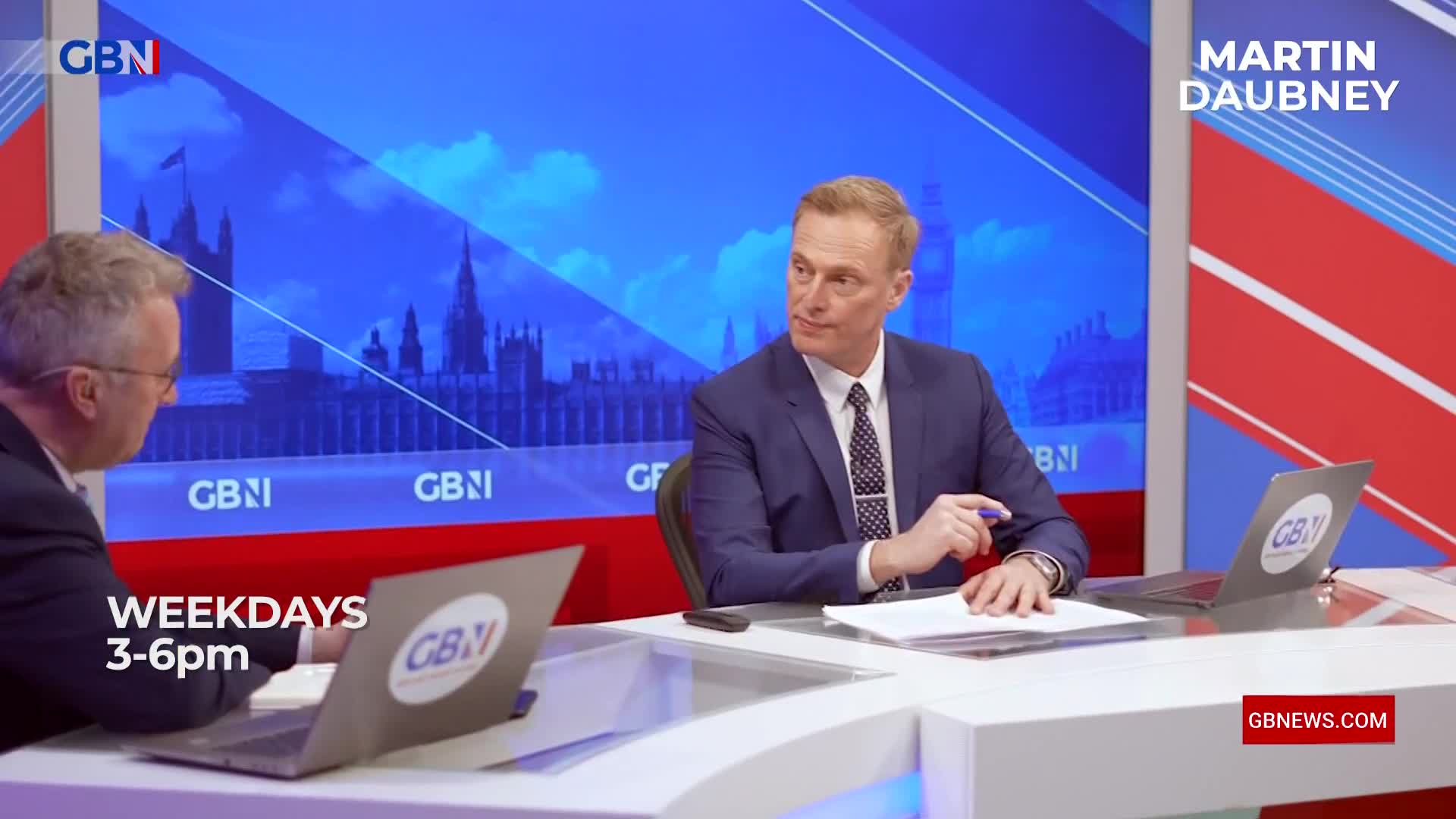 Two men in suits are seated at a desk, with a backdrop of the Houses of Parliament. A laptop with the GB News logo sits open on the desk in front of one of them.