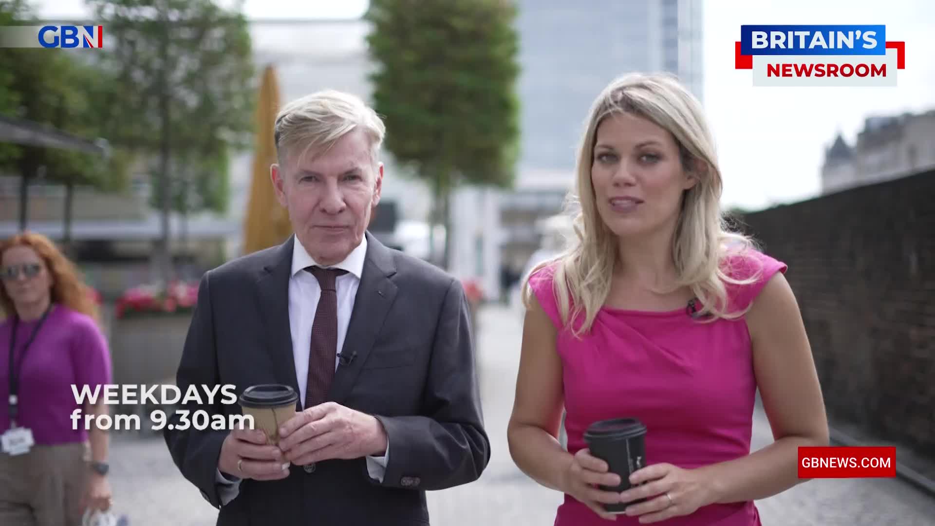 Two presenters from GB News walk towards me, each holding a coffee cup. A woman in a bright pink dress is speaking, while the man beside her, in a suit, looks ahead.