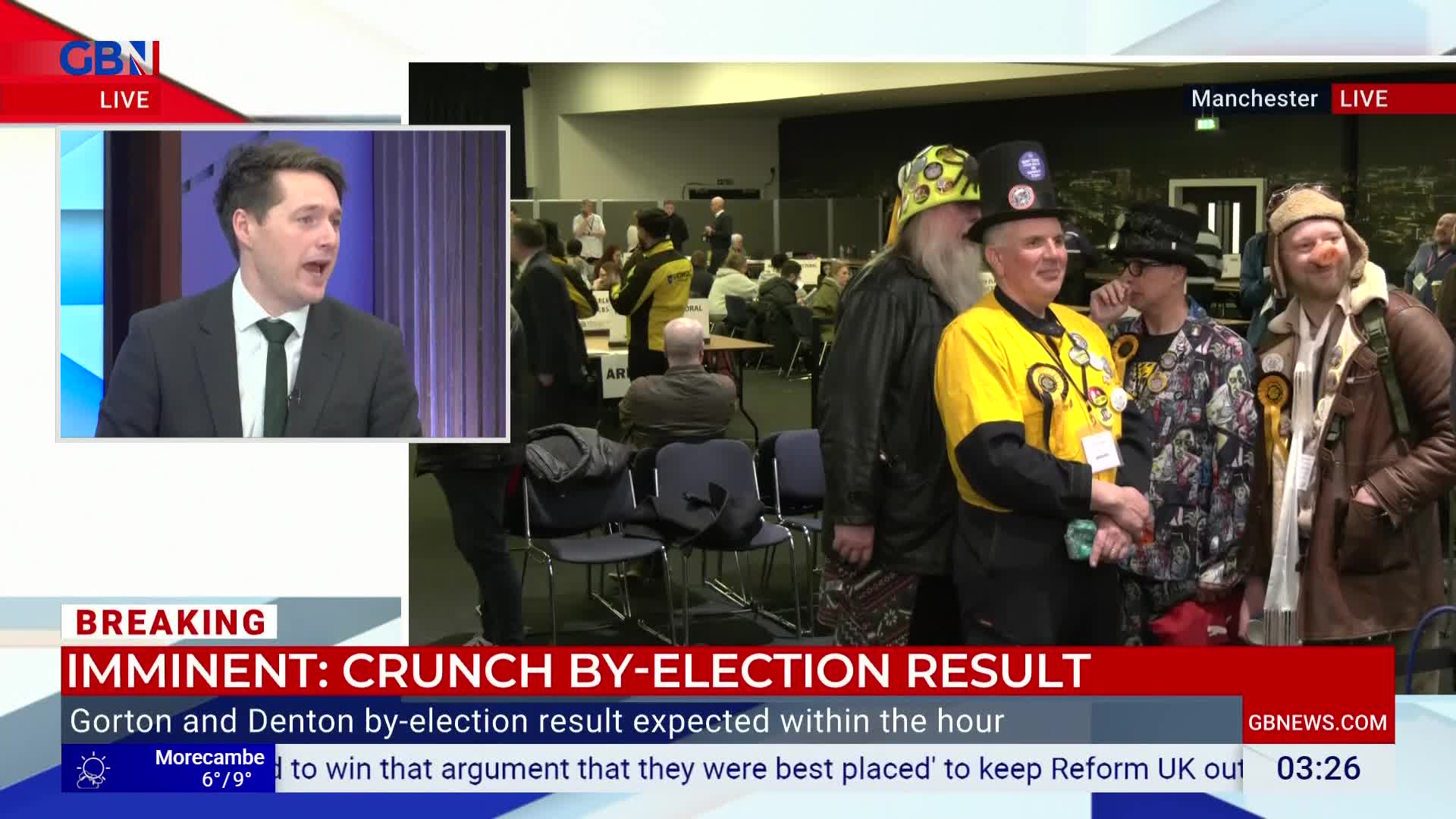 A man in a suit is speaking on GB News, while a group of people in distinctive hats and yellow tops stand nearby. The backdrop appears to be a hall where a by-election result is anticipated.