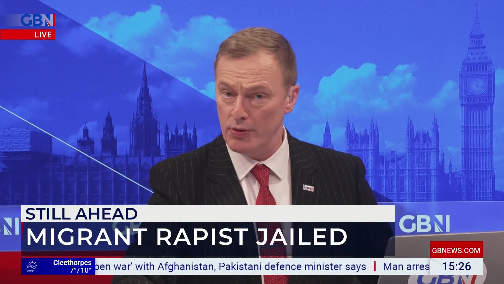 A man in a pinstripe suit speaks directly to the camera, with the Houses of Parliament and Big Ben visible behind him on a screen. A chyron at the bottom reads "MIGRANT RAPIST JAILED."