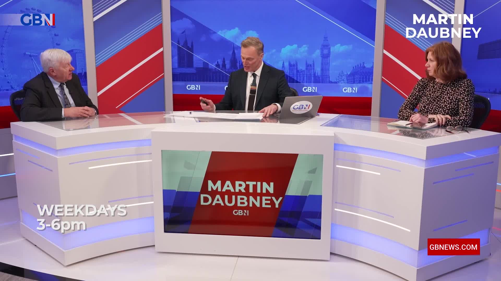 A man in a suit gestures with a pen while speaking to a woman across a desk. Behind them, a graphic displays the Houses of Parliament, a familiar London landmark.