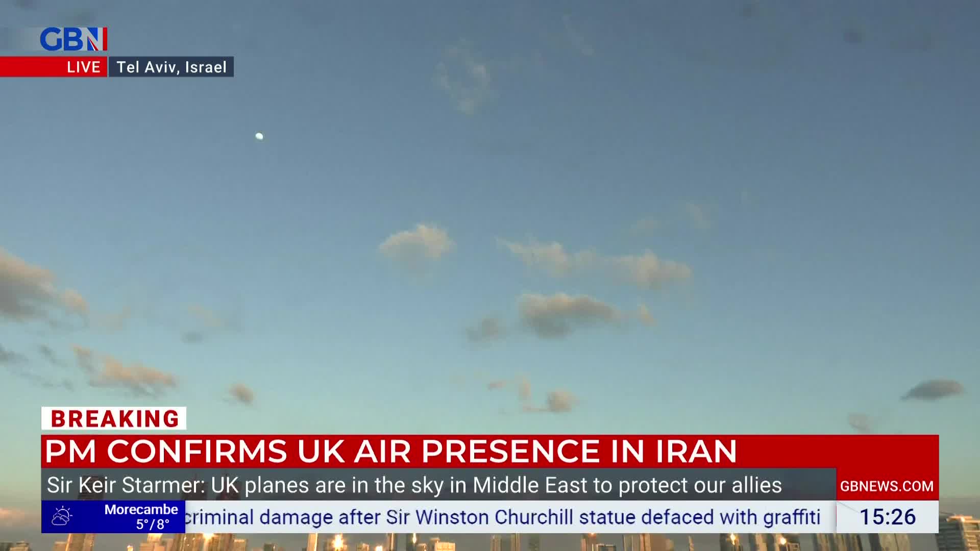 A small, bright object hangs in the pale blue sky. Below, a news ticker scrolls across the screen, reporting on UK air presence in the Middle East.