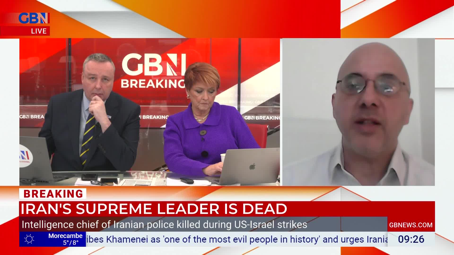 A man with glasses speaks directly to the camera, his face filling the right side of the screen. To his left, two presenters sit at a desk in front of a red backdrop with "GBN BREAKING" displayed prominently. A news ticker scrolls across the bottom, reporting on the death of Iran's Supreme Leader.
