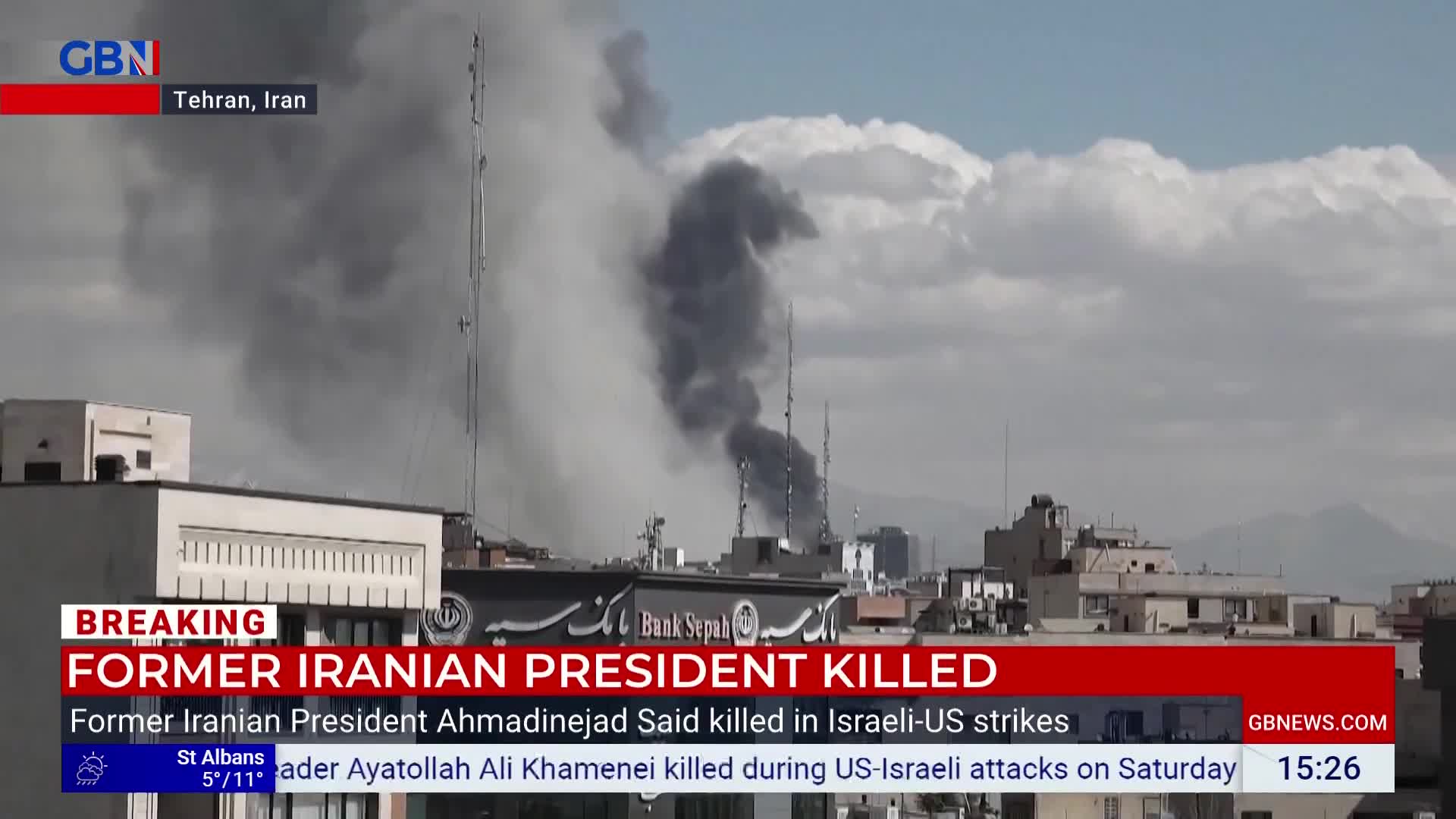 Thick plumes of dark smoke billow into the sky from behind buildings. A news ticker at the bottom of the screen reads "FORMER IRANIAN PRESIDENT KILLED."