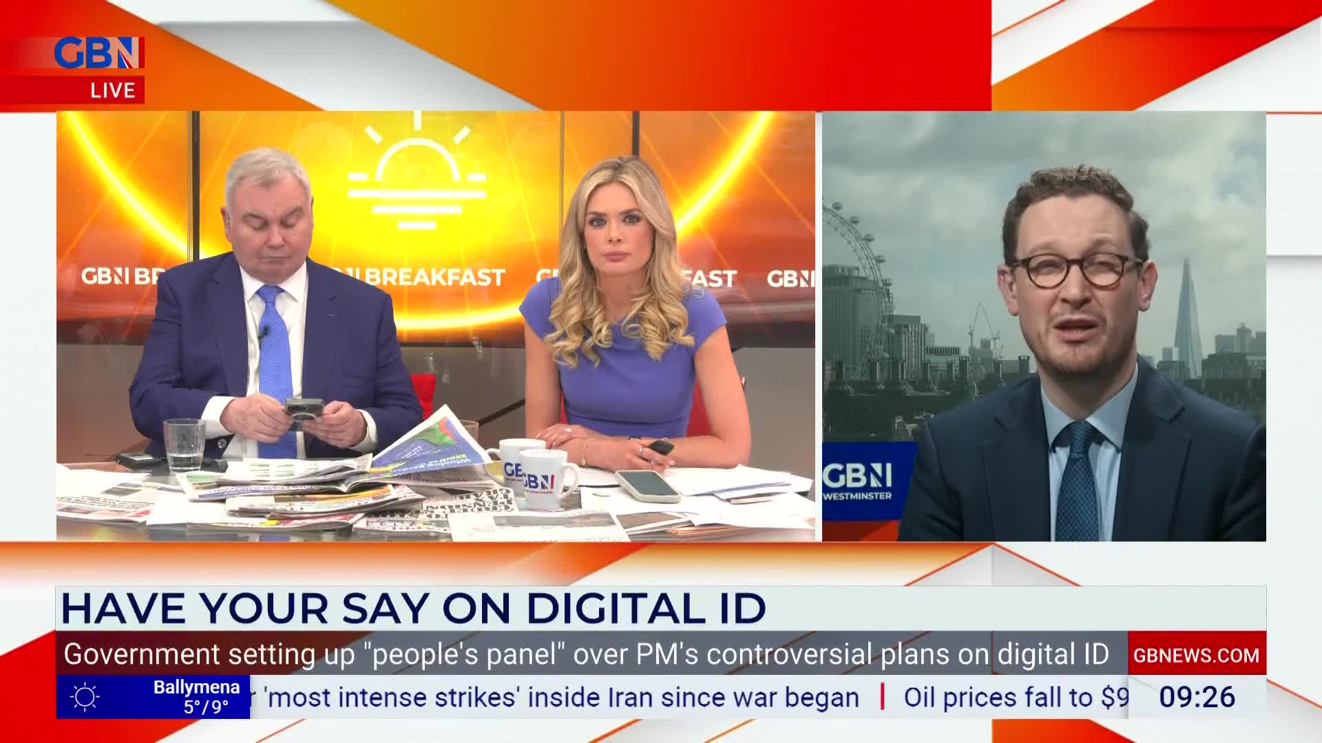 A man in a suit and tie speaks from a screen, his glasses reflecting the light. In front of him, two presenters sit at a desk, one looking down at his phone.