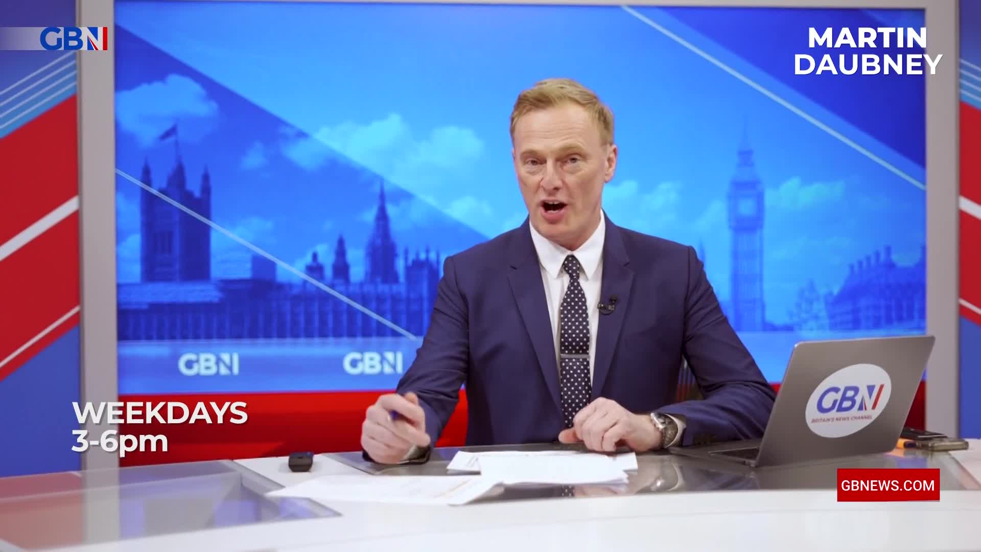 A man in a suit speaks animatedly at a desk, his right hand gesturing forward. Behind him, a screen displays a blue sky over the Houses of Parliament and Big Ben. A man in a suit speaks animatedly at a desk, his right hand gesturing forward. Behind him, a screen displays a blue sky over the Houses of Parliament and Big Ben.