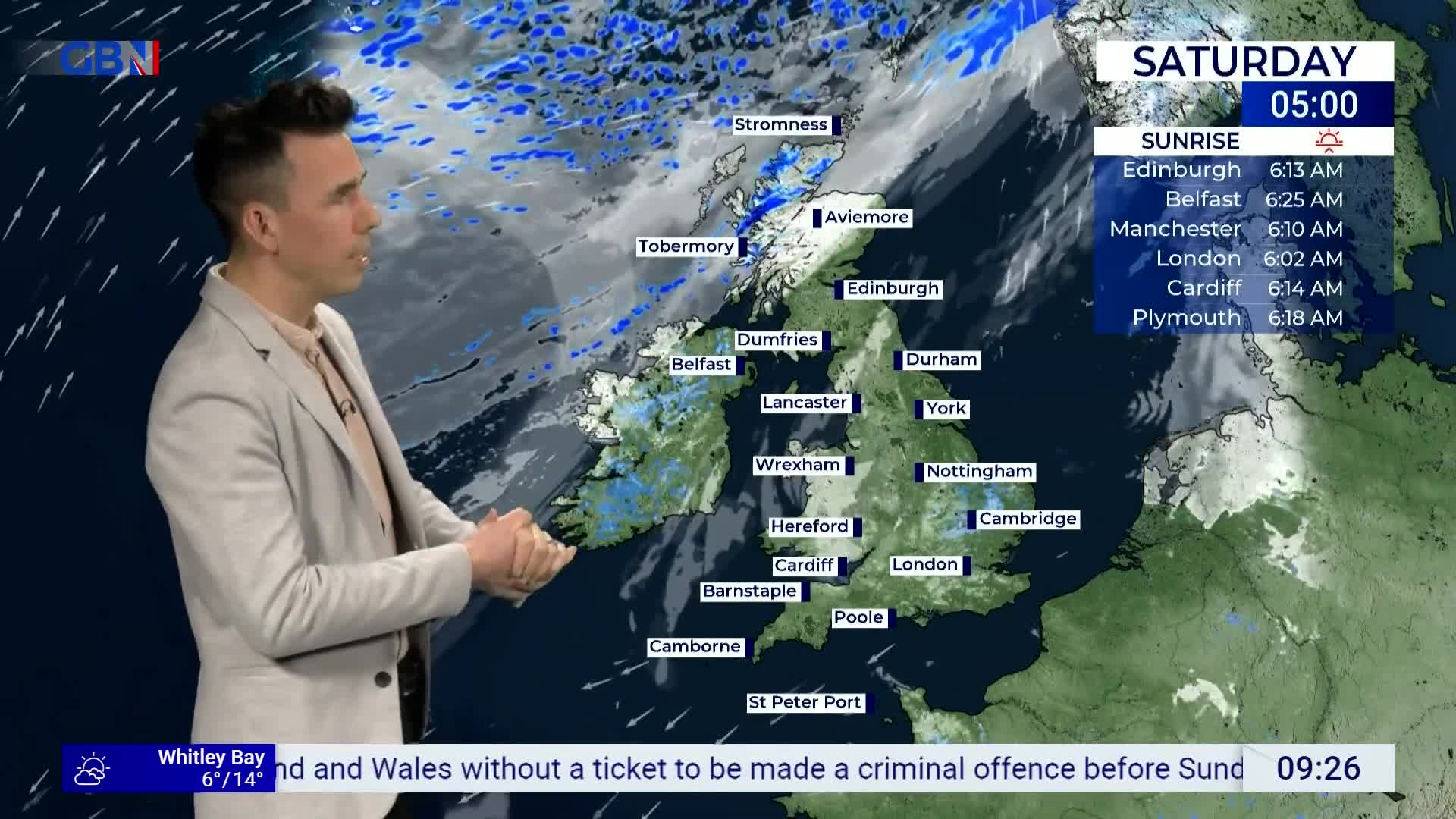A man in a light jacket gestures towards a weather map of the United Kingdom, showing scattered rain across the country. The screen displays sunrise times for several cities, with London's at 6:02 AM. A man in a light jacket gestures towards a weather map of the United Kingdom, showing scattered rain across the country. The screen displays sunrise times for several cities, with London's at 6:02 AM.