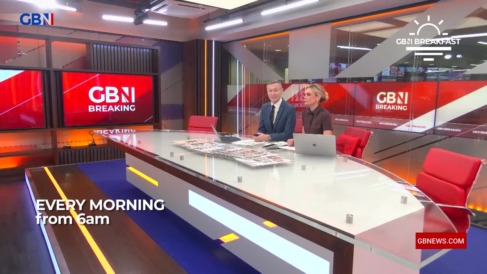 Two presenters sit at a large desk in a modern studio, surrounded by screens displaying the GB News logo. Newspapers are spread across the desk between them. Two presenters sit at a large desk in a modern studio, surrounded by screens displaying the GB News logo. Newspapers are spread across the desk between them.