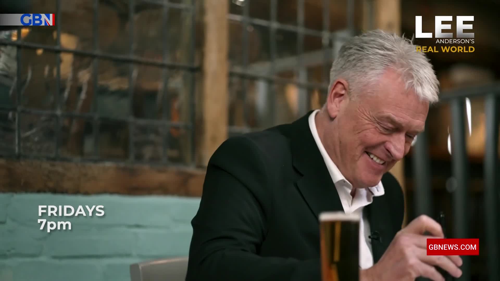 A man with grey hair, dressed in a black suit jacket and white shirt, laughs heartily. A pint of beer sits on the table in front of him. A man with grey hair, dressed in a black suit jacket and white shirt, laughs heartily. A pint of beer sits on the table in front of him.