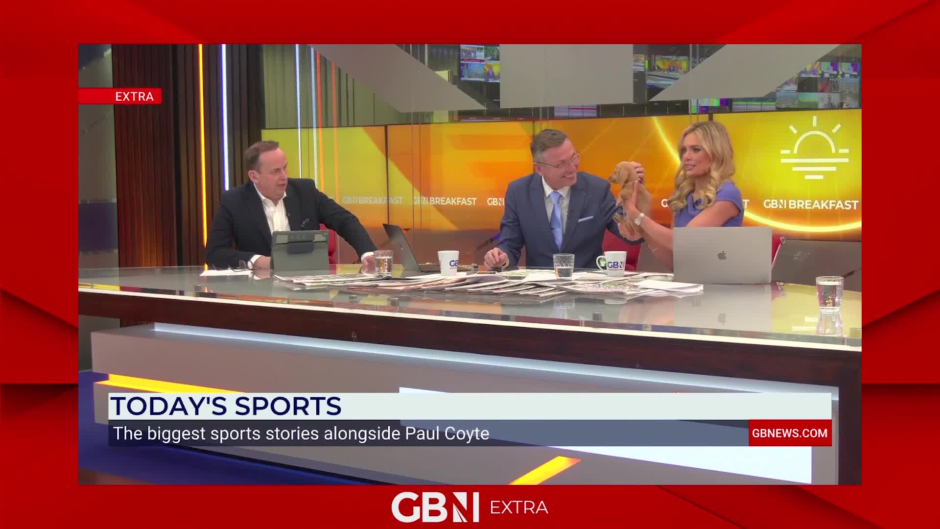 A man in a suit is holding a small, light brown dog up to a woman on the other side of the desk. Newspapers are spread across the table in front of them. A man in a suit is holding a small, light brown dog up to a woman on the other side of the desk. Newspapers are spread across the table in front of them.