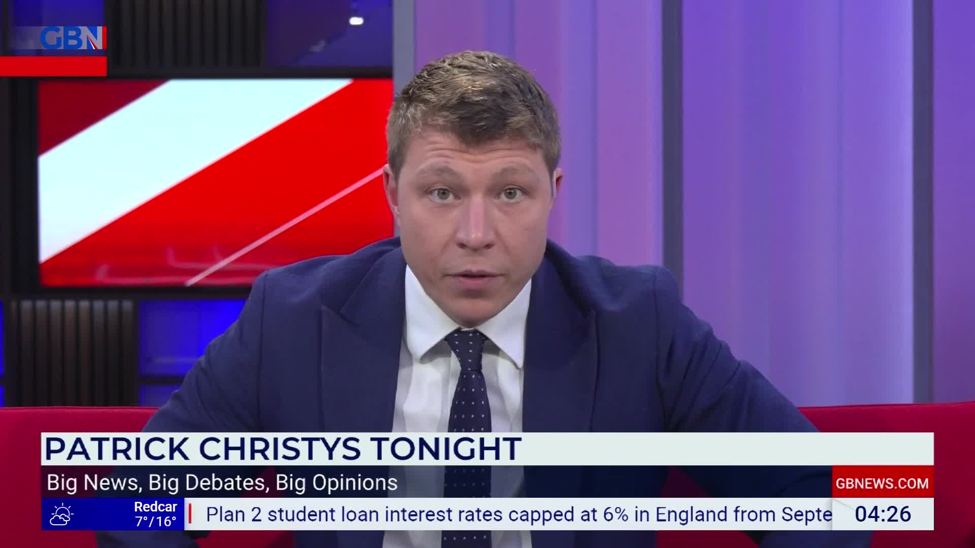 A man in a suit and tie sits on a red sofa, looking directly ahead. Behind him, a screen displays the GB News logo and a red and white graphic. A ticker at the bottom of the screen shows news headlines and the current time. A man in a suit and tie sits on a red sofa, looking directly ahead. Behind him, a screen displays the GB News logo and a red and white graphic. A ticker at the bottom of the screen shows news headlines and the current time.