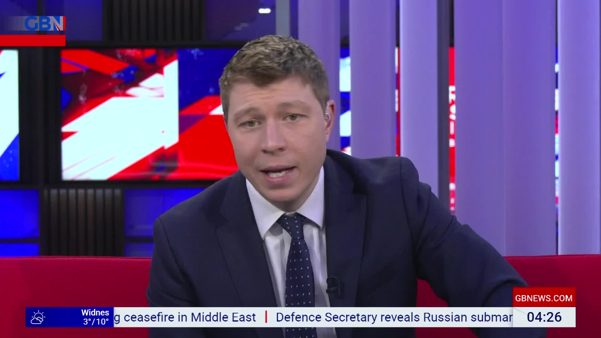 A man in a suit speaks directly to the camera, with a red sofa behind him. A ticker tape displaying news headlines scrolls across the bottom of the screen. A man in a suit speaks directly to the camera, with a red sofa behind him. A ticker tape displaying news headlines scrolls across the bottom of the screen.