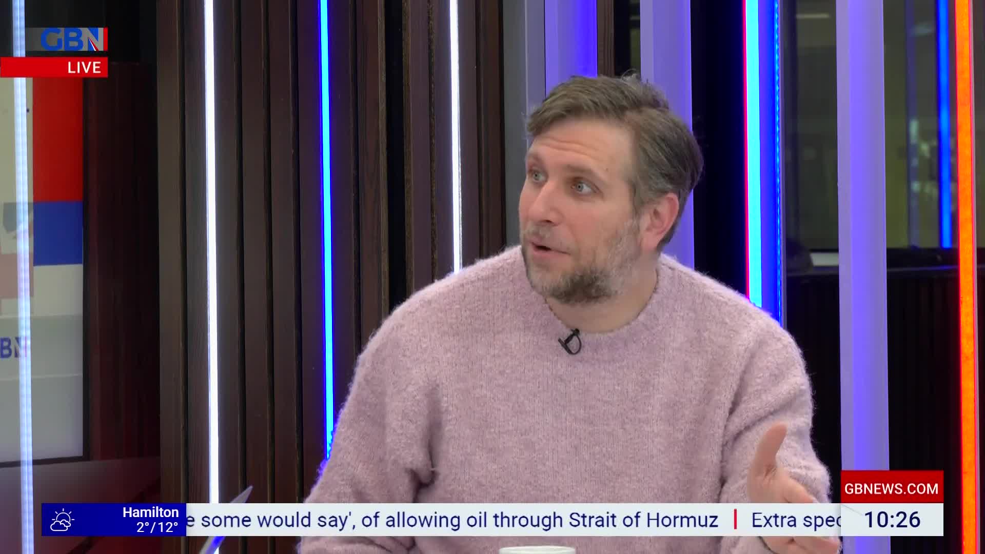 A man in a pink sweater is speaking animatedly. Behind him, vertical lights glow blue and white. A man in a pink sweater is speaking animatedly. Behind him, vertical lights glow blue and white.