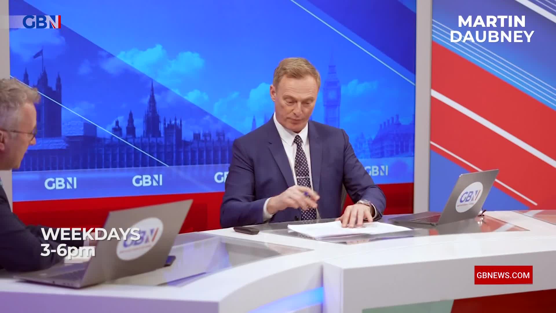 A man in a suit leans forward, pen in hand, over papers on a desk. Behind him, a screen displays the Houses of Parliament and the GB News logo. A man in a suit leans forward, pen in hand, over papers on a desk. Behind him, a screen displays the Houses of Parliament and the GB News logo.