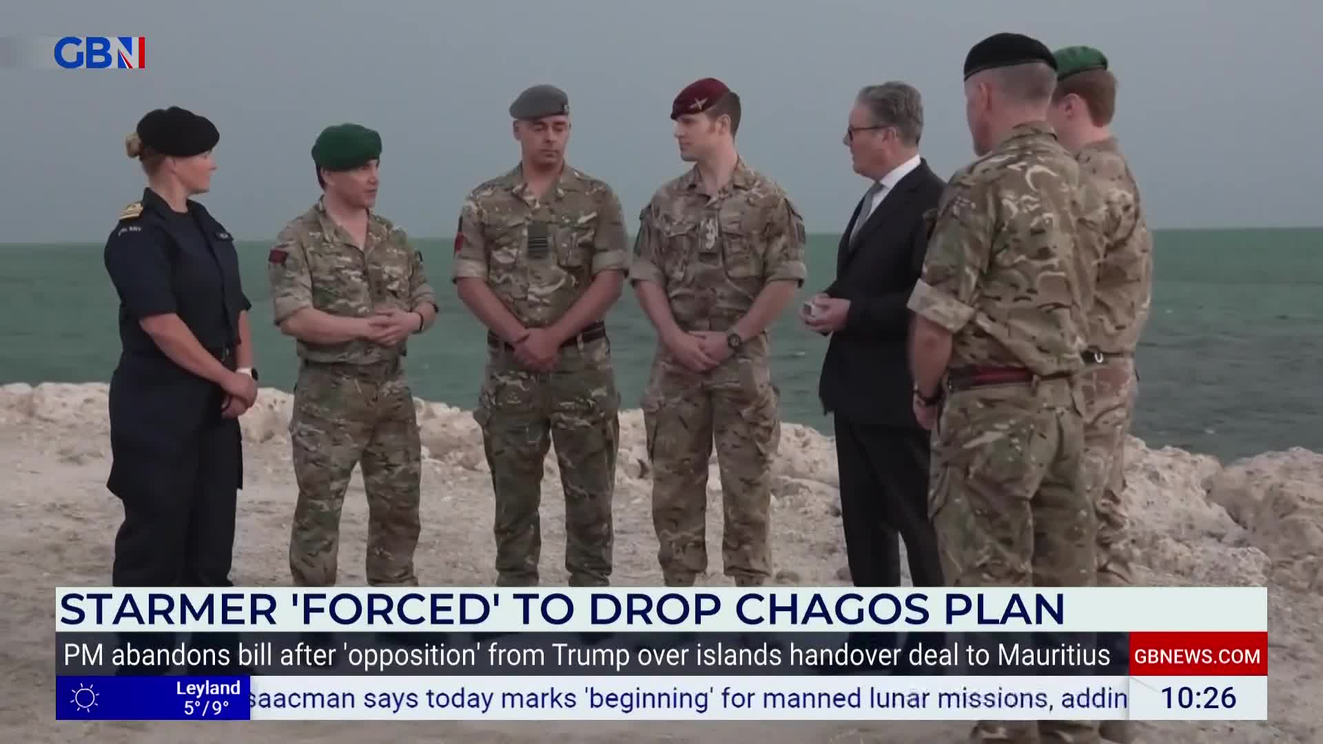 A group of British military personnel stand on a rocky shore, facing a man in a suit. The sea stretches out behind them under a grey sky. A group of British military personnel stand on a rocky shore, facing a man in a suit. The sea stretches out behind them under a grey sky.