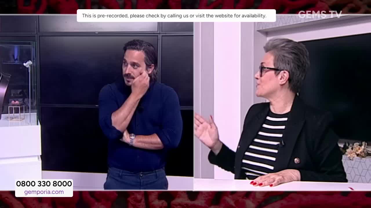 The man in the blue sweater is resting his hand on his cheek, while the woman in the striped shirt gestures with her hand. They're on a Gems TV set, discussing something, with a display case of jewelry off to the side.
