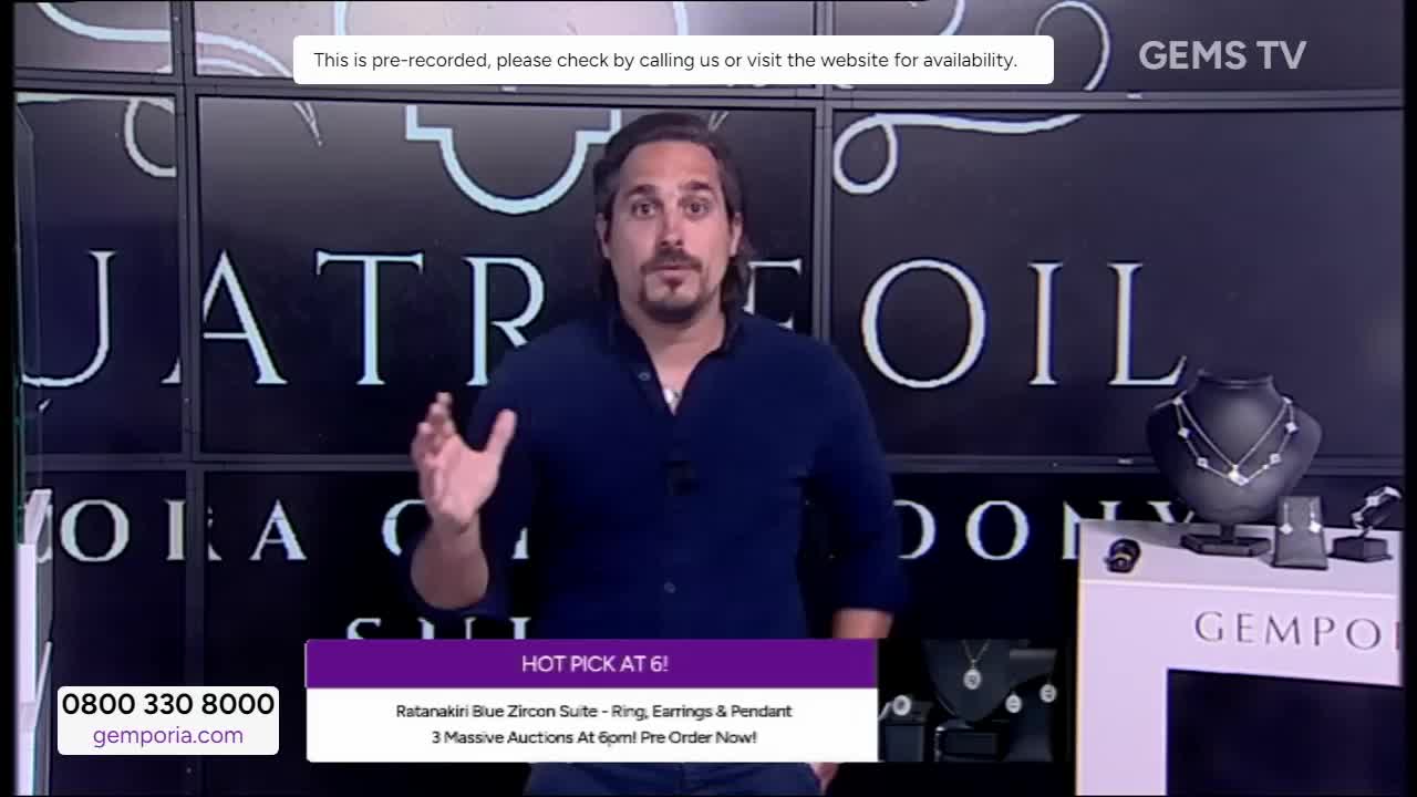 The man in the blue shirt gestures with both hands as he speaks, likely on Gems TV in the United Kingdom. Behind him, jewelry is displayed on stands, and a graphic advertises a "Hot Pick" for sale.
