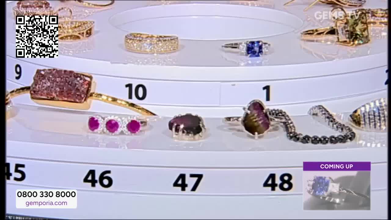 A rotating display of jewelry is set up, with each piece numbered and lit. The Gems TV logo is visible, and a "Coming Up" preview of a ring with a blue stone appears at the bottom.
