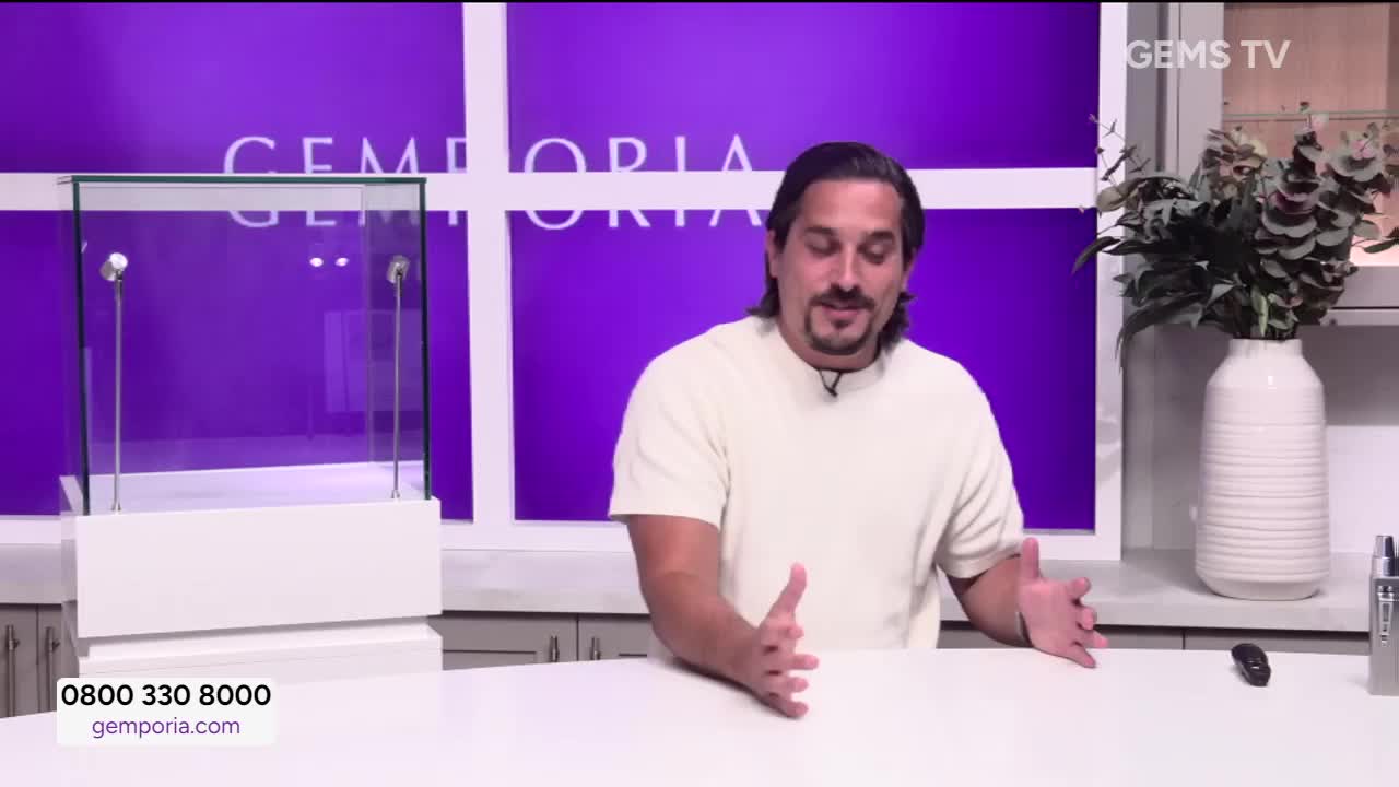 A man in a cream-colored shirt gestures with his hands, speaking to the camera. Behind him, a display case sits on a white counter, ready for the jewelry.
