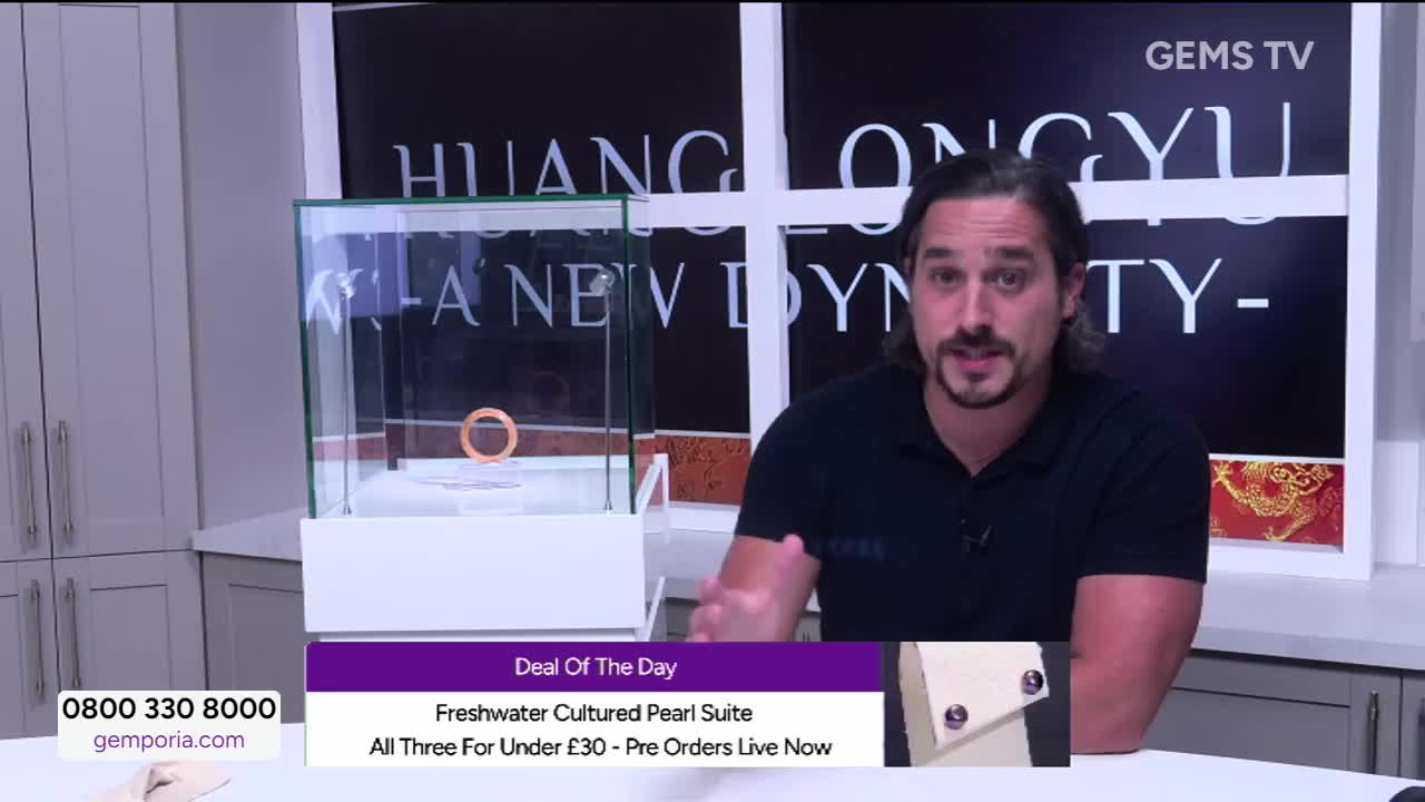 A man in a black shirt gestures towards a glass display case, where a ring sits on a white platform. The Gems TV logo is visible in the corner, and a "Deal of the Day" banner is displayed on the screen.
