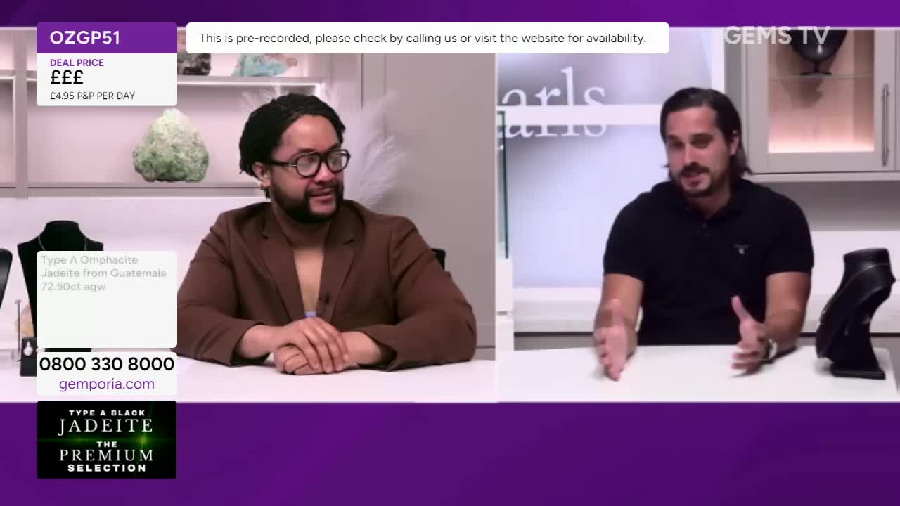 Two men sit at a white table, talking on a Gems TV broadcast. One gestures with his hands, while the other looks on, a display of jewelry visible in the foreground.
