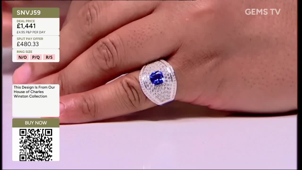 A hand, likely on a Gems TV broadcast, is displaying a large, ornate ring. The ring features a central blue stone surrounded by smaller, glittering gems.
