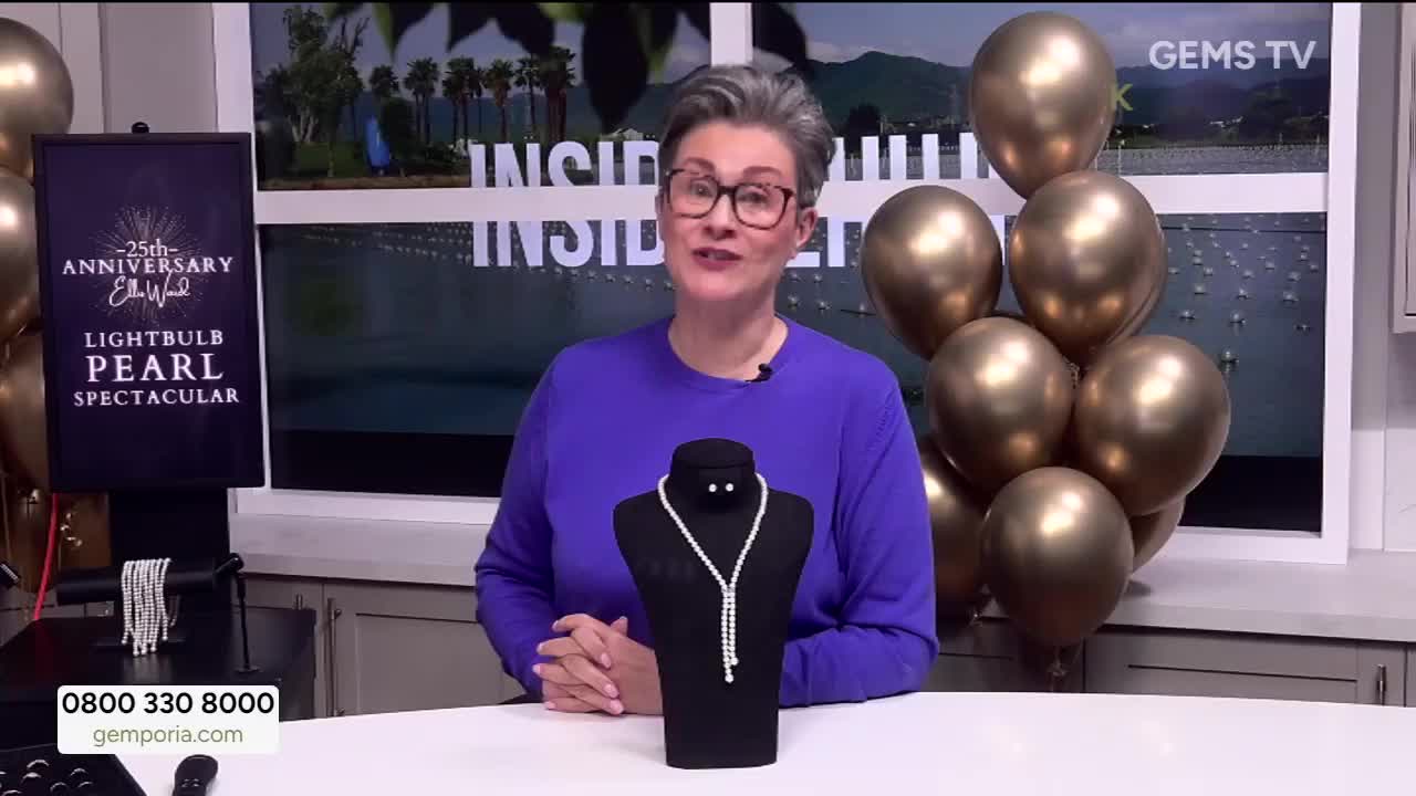 A woman in a purple sweater stands in front of a black display, gesturing towards a pearl necklace. Behind her, a cluster of gold balloons and a sign for a "Lightbulb Pearl Spectacular" are visible.
