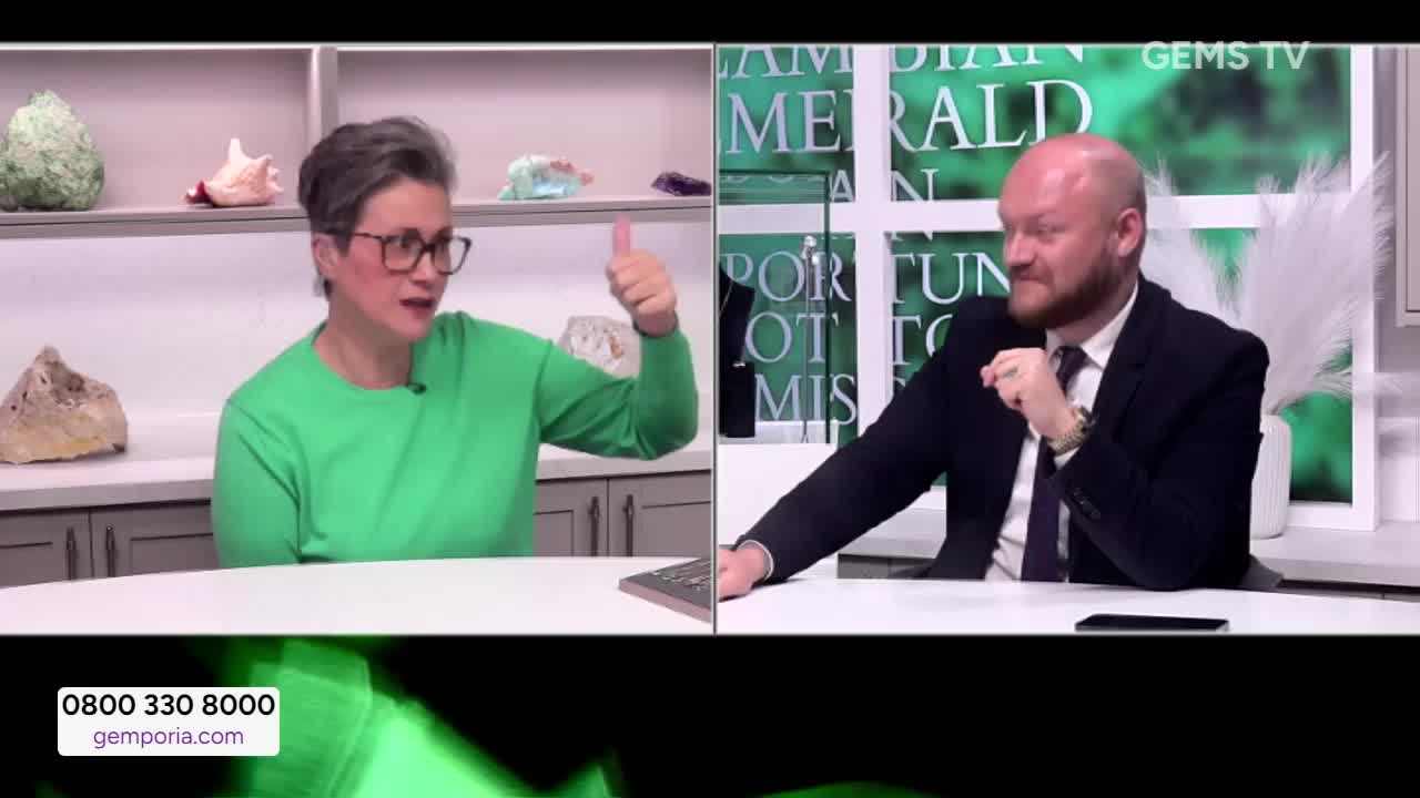 The woman in the green sweater is giving a thumbs-up while talking, and the man in the suit is listening intently. Behind them, the Gems TV logo is visible.
