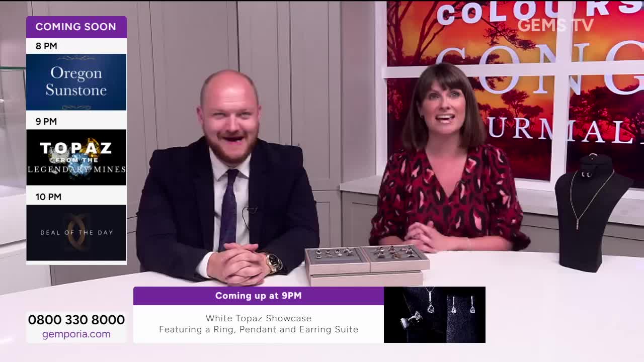 The two presenters on Gems TV are smiling, looking directly at the camera. A display of jewelry sits in front of them, and a graphic lists the upcoming shows.
